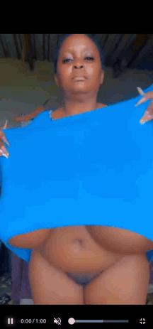 Busty mature ebony woman in a blue shirt and thong performing a titty drop in her bedroom.
