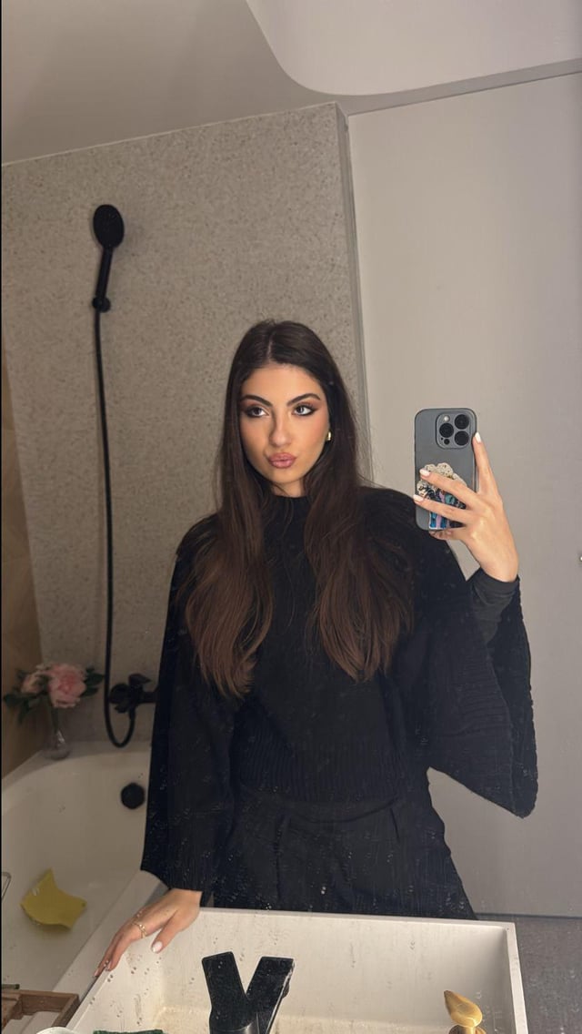 Slim brunette woman in a black long-sleeved top and skirt taking a mirror selfie in a bathroom
