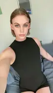 Androgynous woman with short brown hair in a black sleeveless bodysuit posing for a selfie in a bedroom