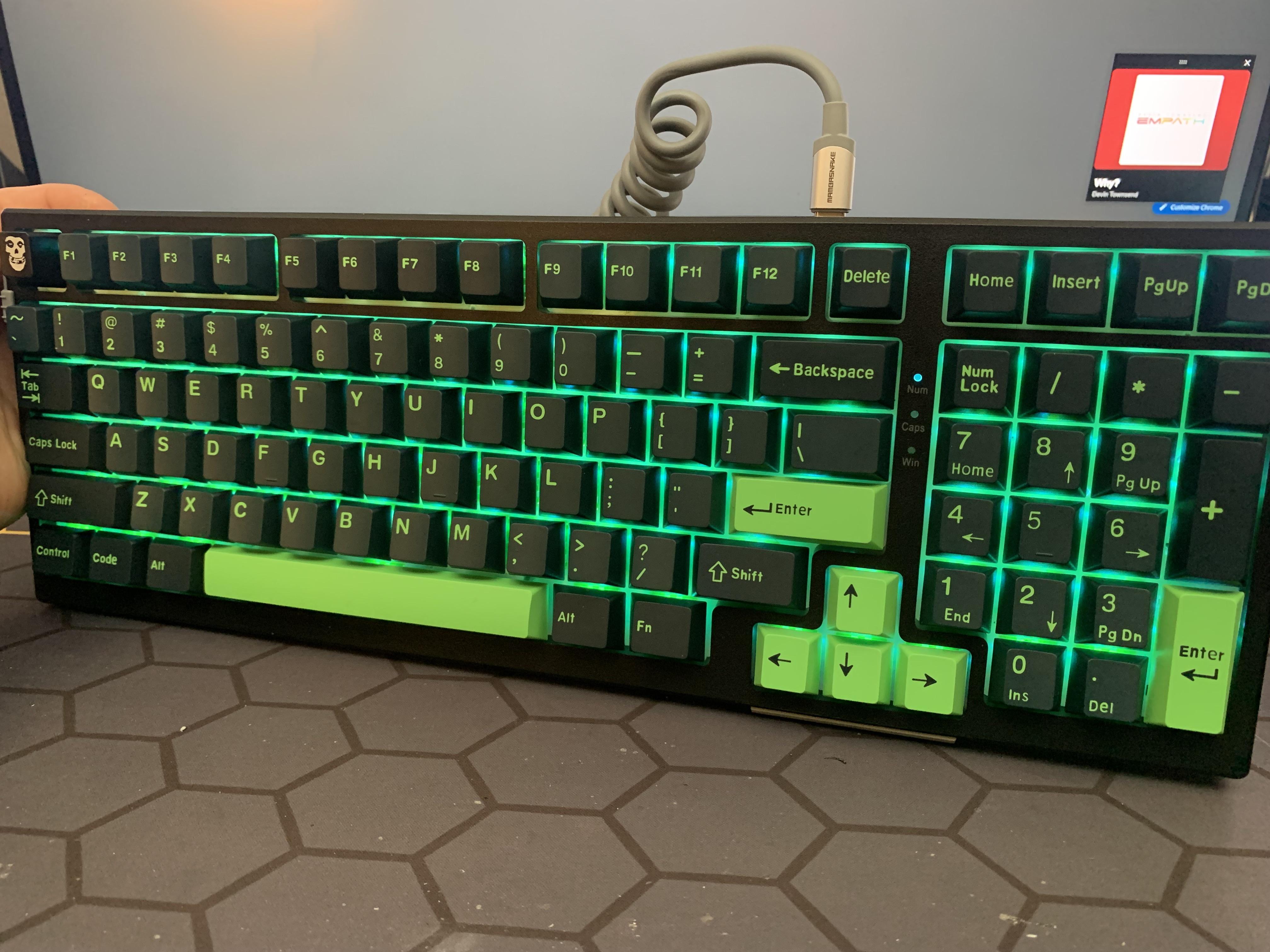 Close-up of a MageGee Sky98 mechanical keyboard with green backlit keys and a coiled cable on a grey hexagonal surface.
