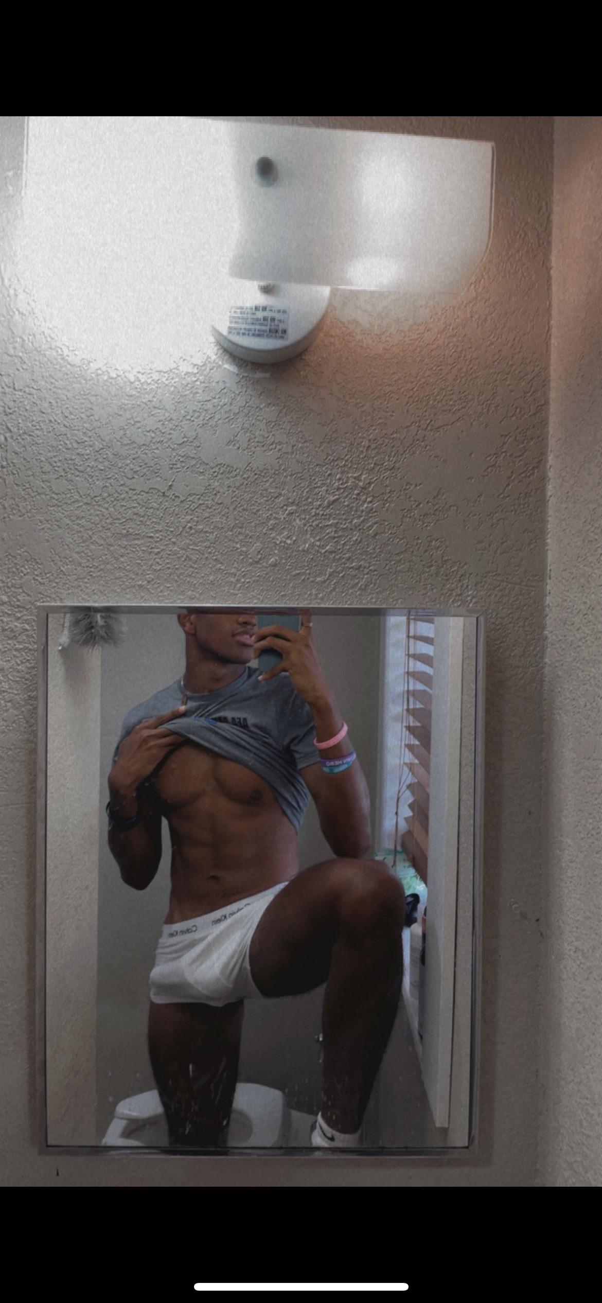 Muscular black man in white Calvin Klein underwear taking a mirror selfie in a bathroom, lifting his grey t-shirt to show abs