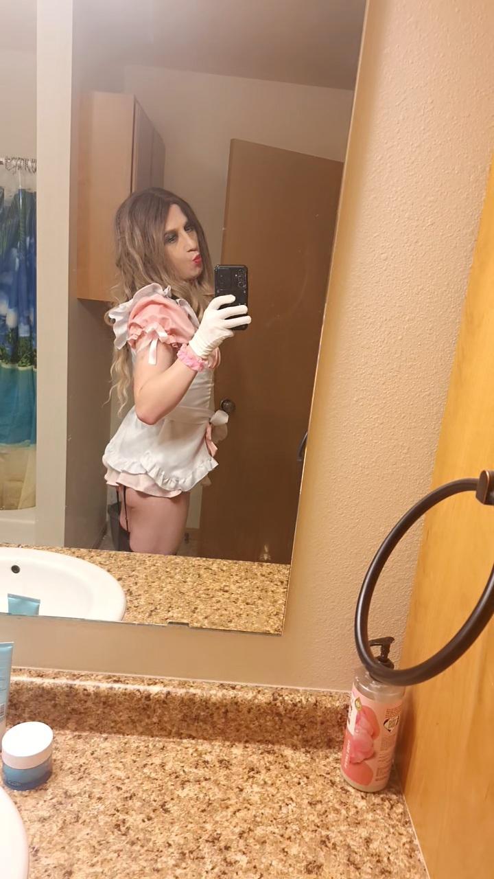 Femboy in a pink maid dress, white gloves, and checkered stockings posing for selfies with a long brown wig.