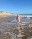 Naked mature white man standing in the ocean waves on a sandy beach for international skinny dipping day.