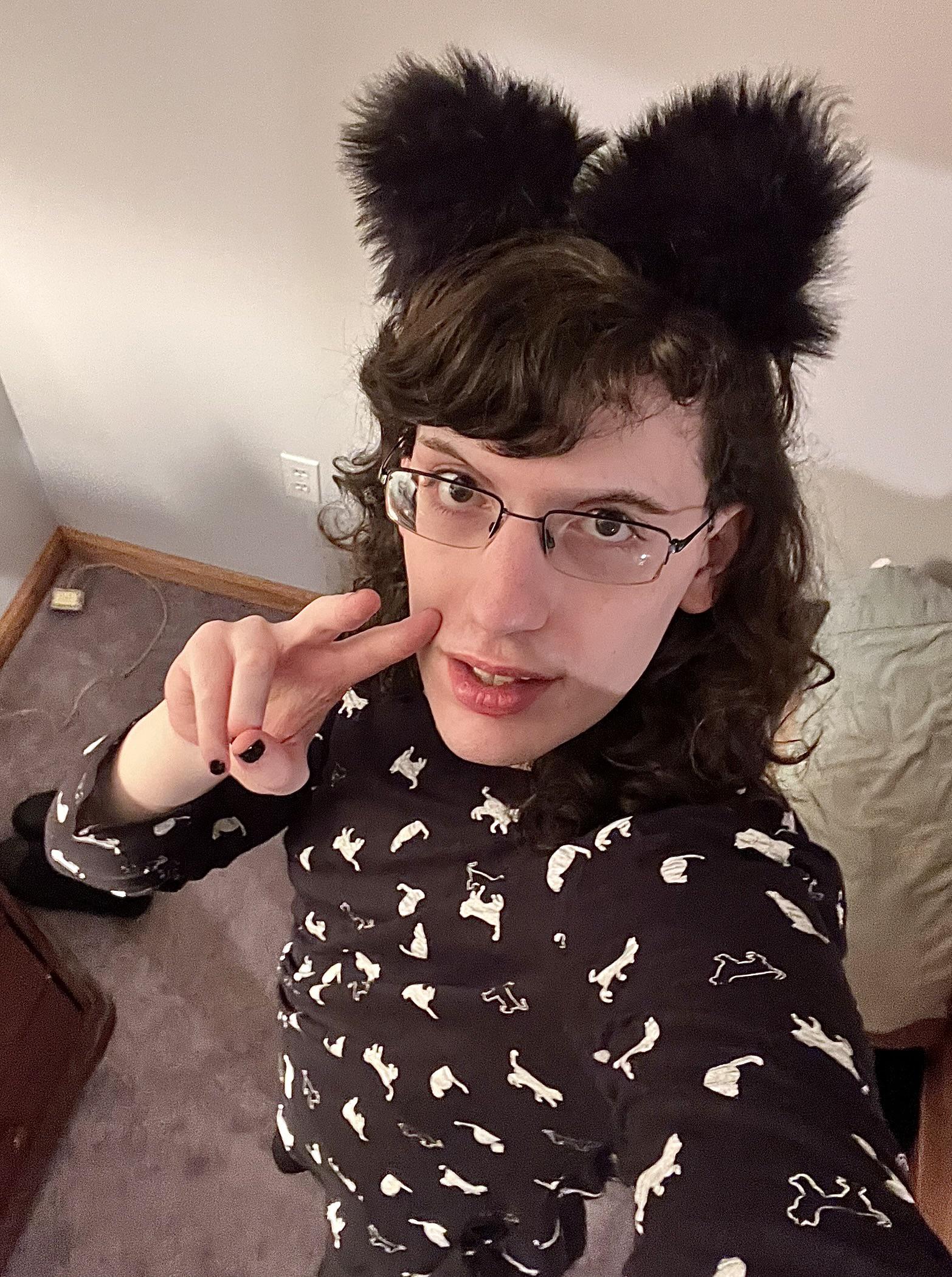 A trans woman with dark curly hair and glasses wearing black fuzzy animal ears and a patterned shirt.