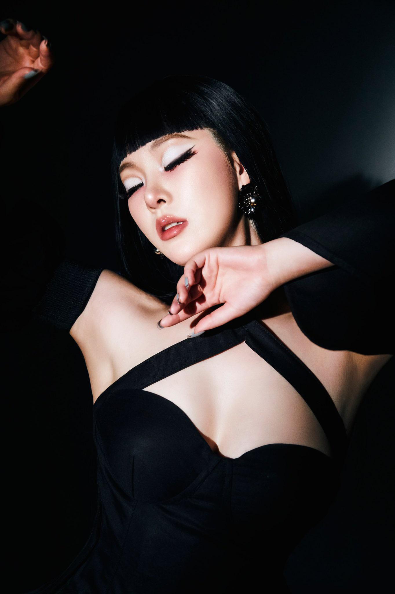 SAAY in a black off-the-shoulder dress with bold eyeliner and black hair with bangs against a dark background