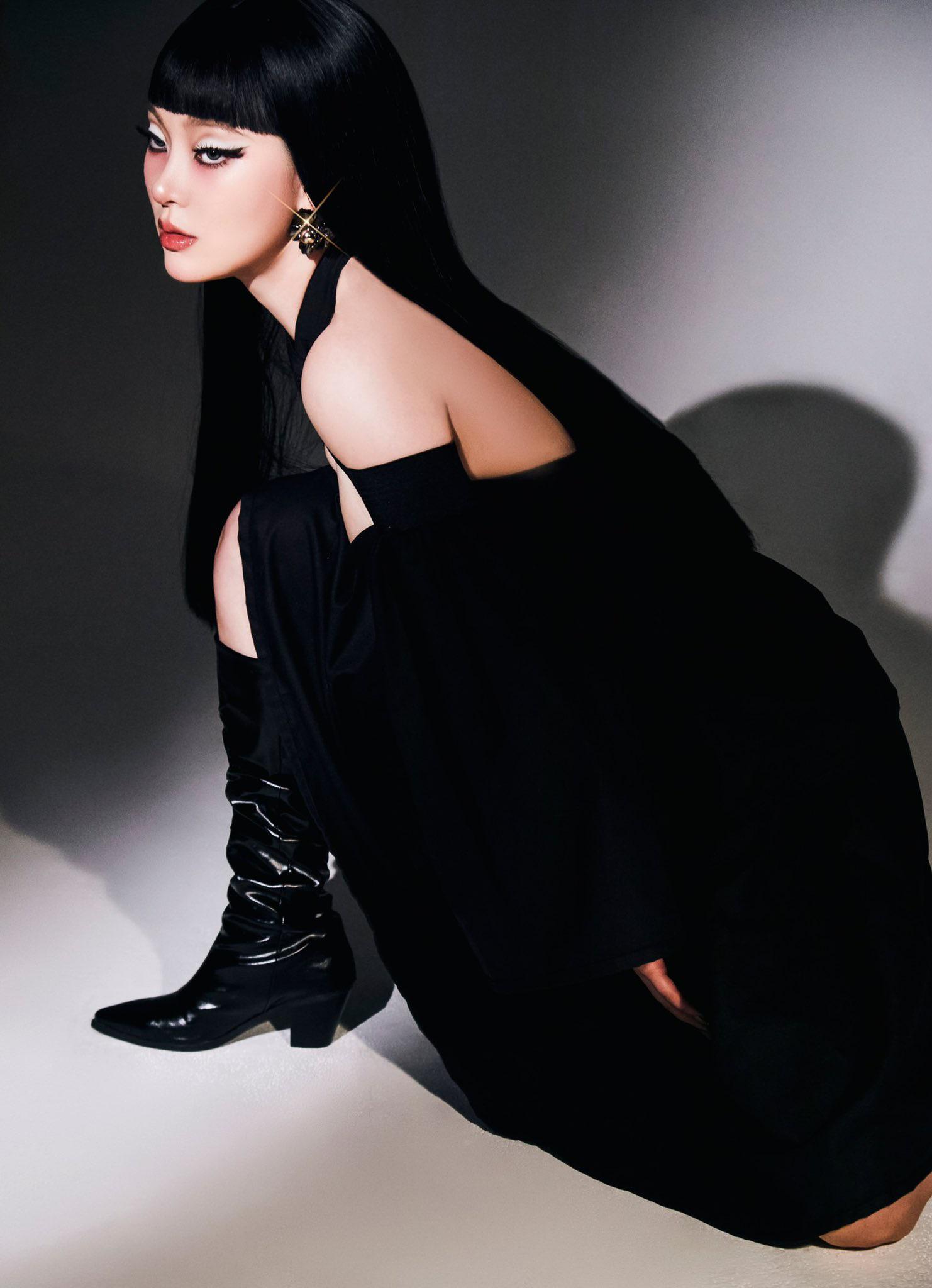 SAAY in a black off-the-shoulder dress with bold eyeliner and black hair with bangs against a dark background
