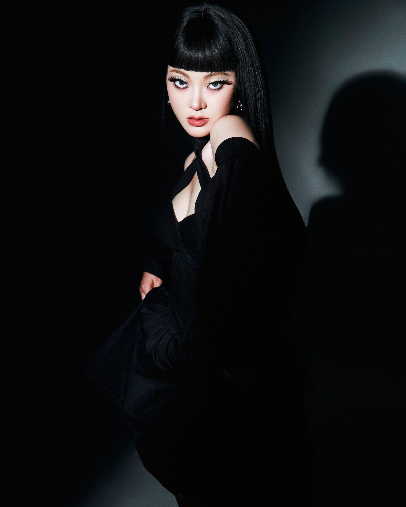 SAAY in a black off-the-shoulder dress with bold eyeliner and black hair with bangs against a dark background