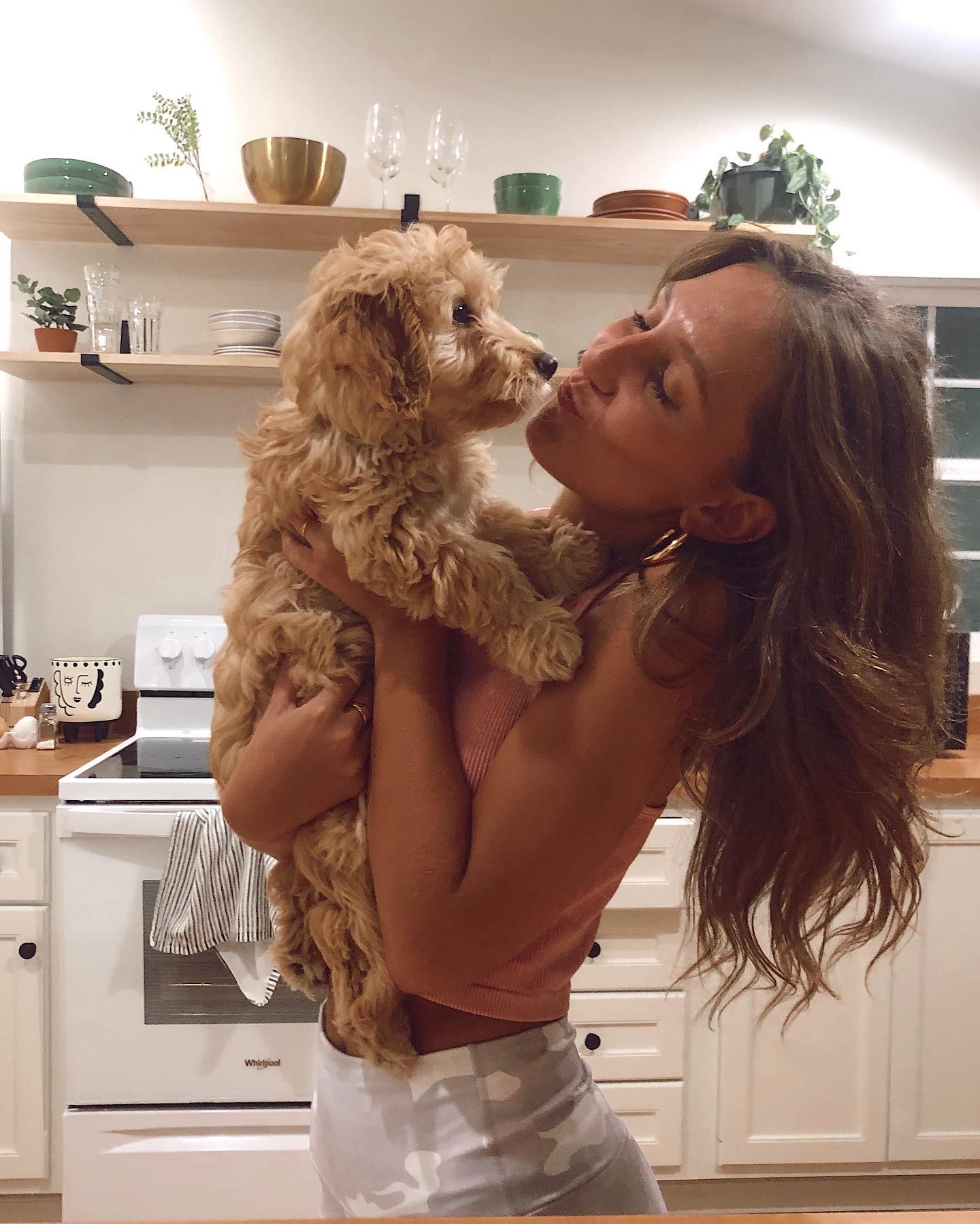 A woman with long brown hair in a pink ribbed top and white skirt holding a fluffy brown dog for doggy kisses.