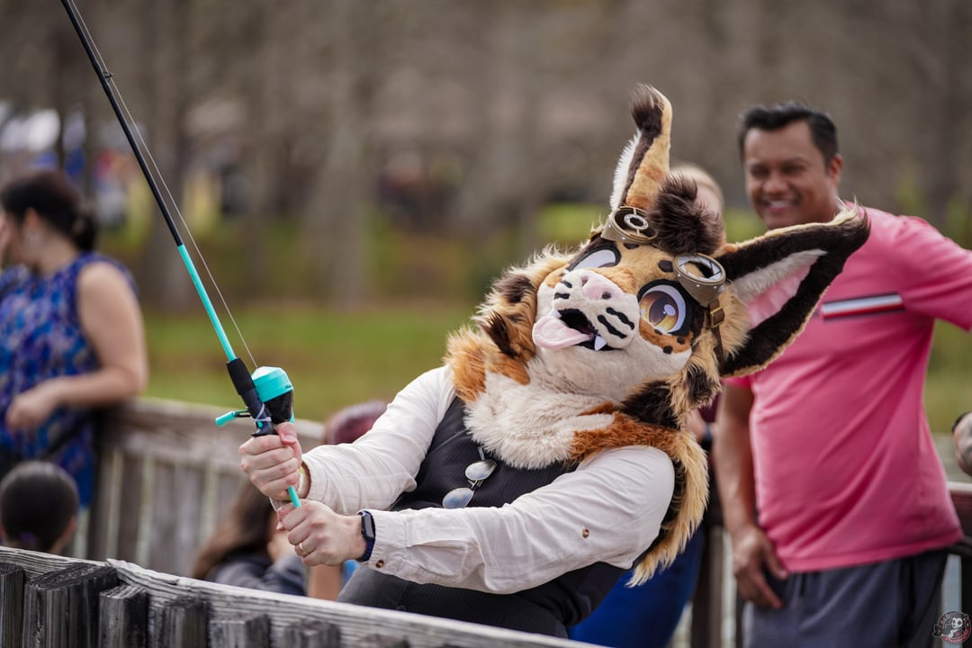 A person in a brown and white animal fursuit with goggles holding a fishing rod outdoors with a group of friends