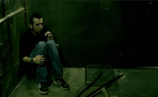 A slender man with pale skin in a black shirt and blue jeans sitting against a wall in a dark, green-tinted room.