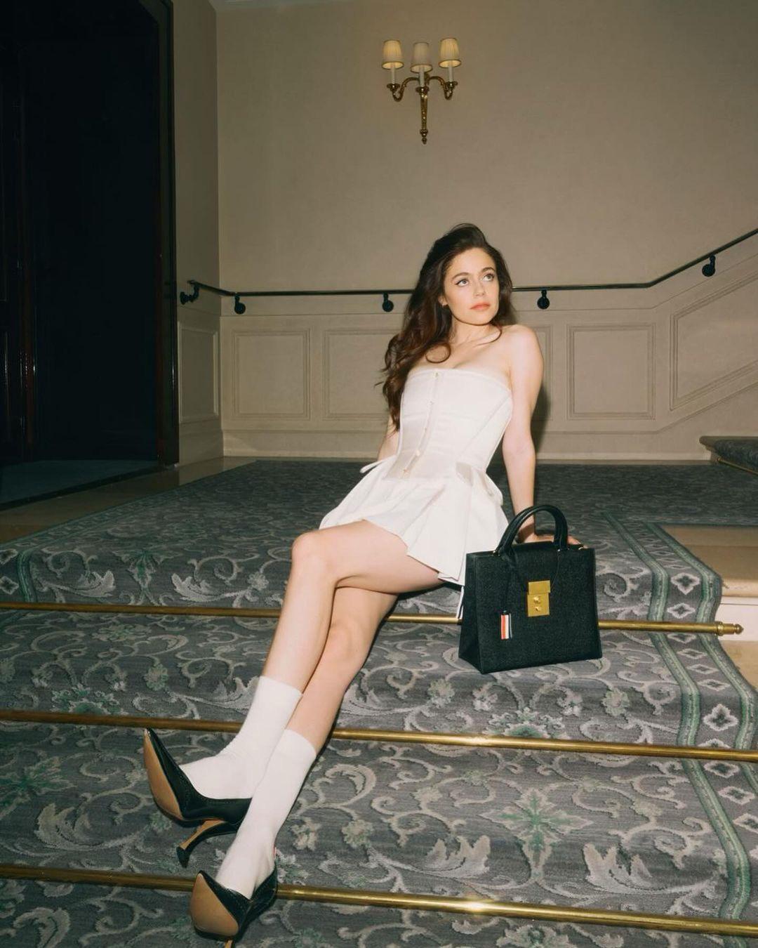 Molly Gordon in a white mini corset dress, white socks, and black heels posing by an ornate staircase