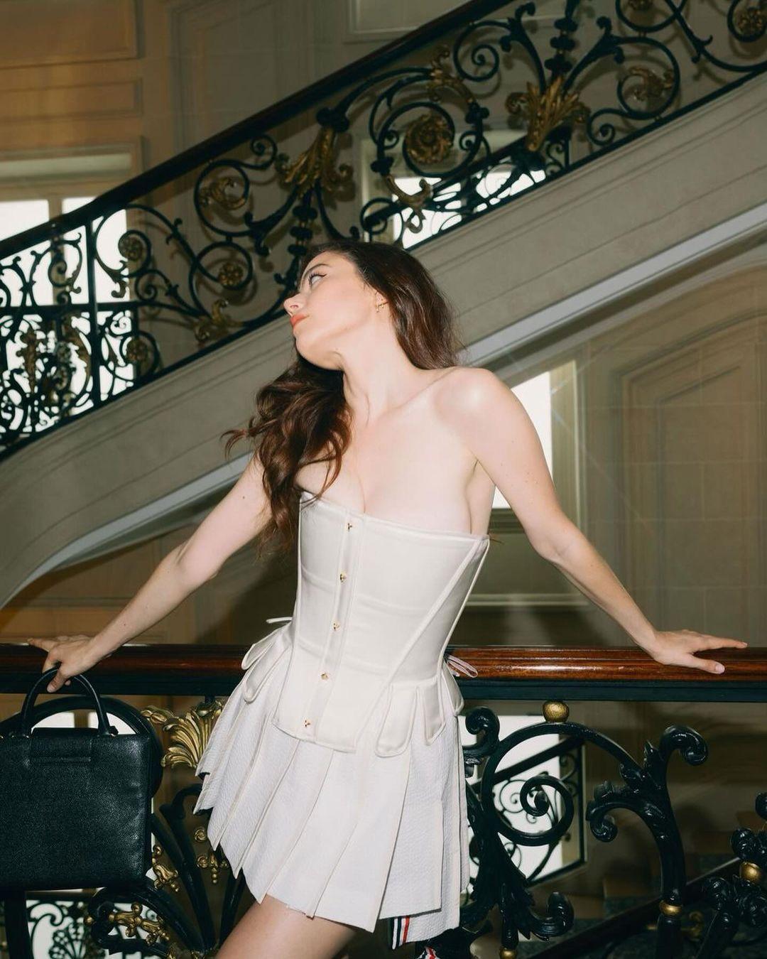 Molly Gordon in a white mini corset dress, white socks, and black heels posing by an ornate staircase