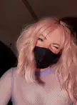 A femboy with pink hair wearing a black face mask and purple mesh top winking for a selfie.