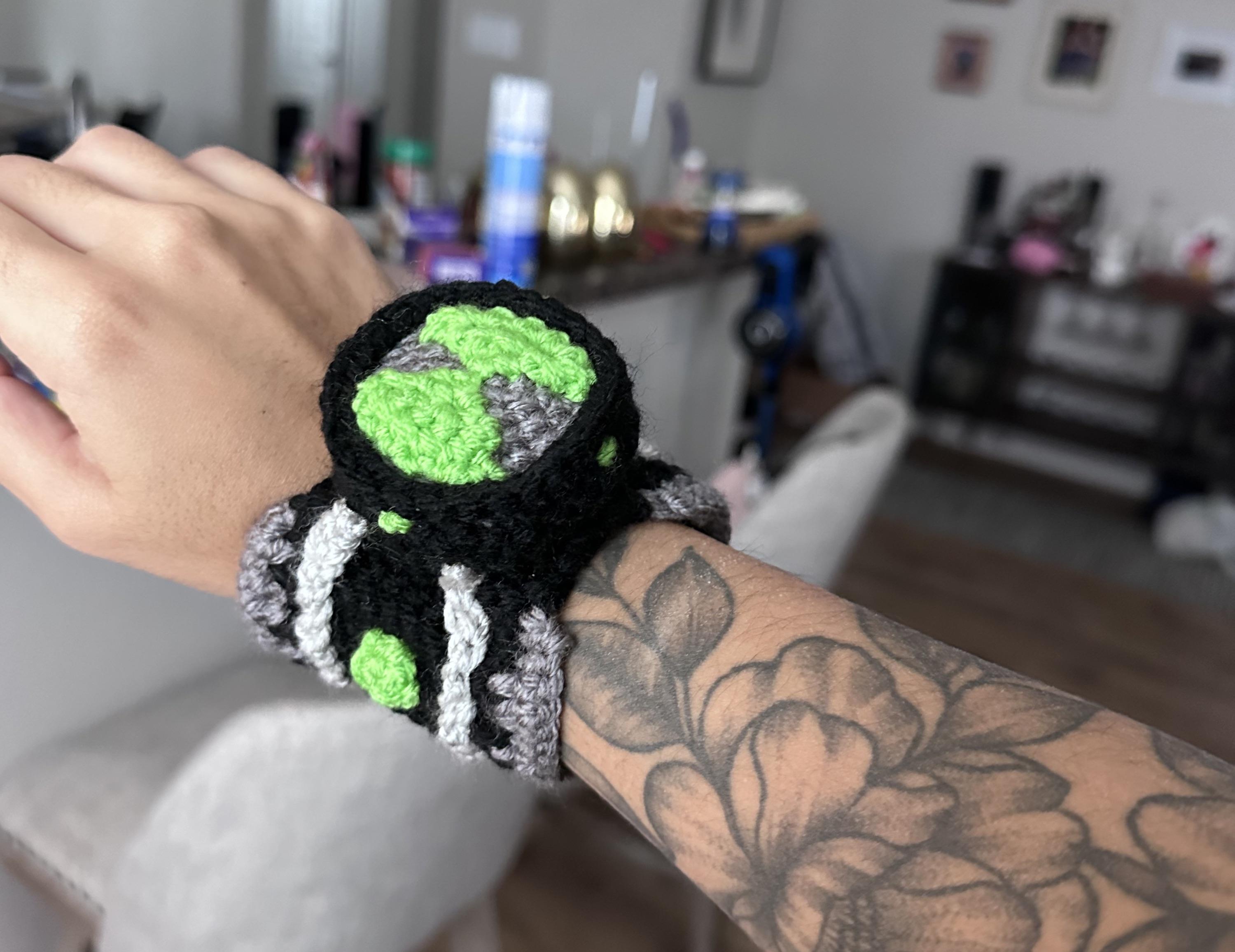 Close-up of a crocheted Omnitrix on a wrist with a fine line floral tattoo on the forearm.