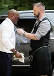 A man in a green and blue plaid kilt and black vest helps a man in a white shirt tie a light blue tie.