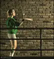 Slender blonde woman in a green jacket and white shorts practicing strikes against a brick wall with a stick