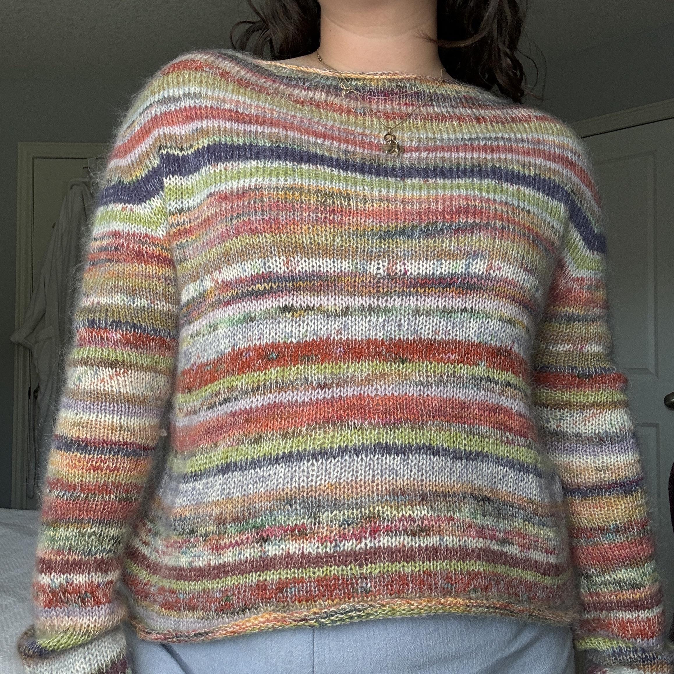 Mirror selfie of a woman with brown hair wearing a colorful, multi-striped mini skein knitted sweater and blue jeans.