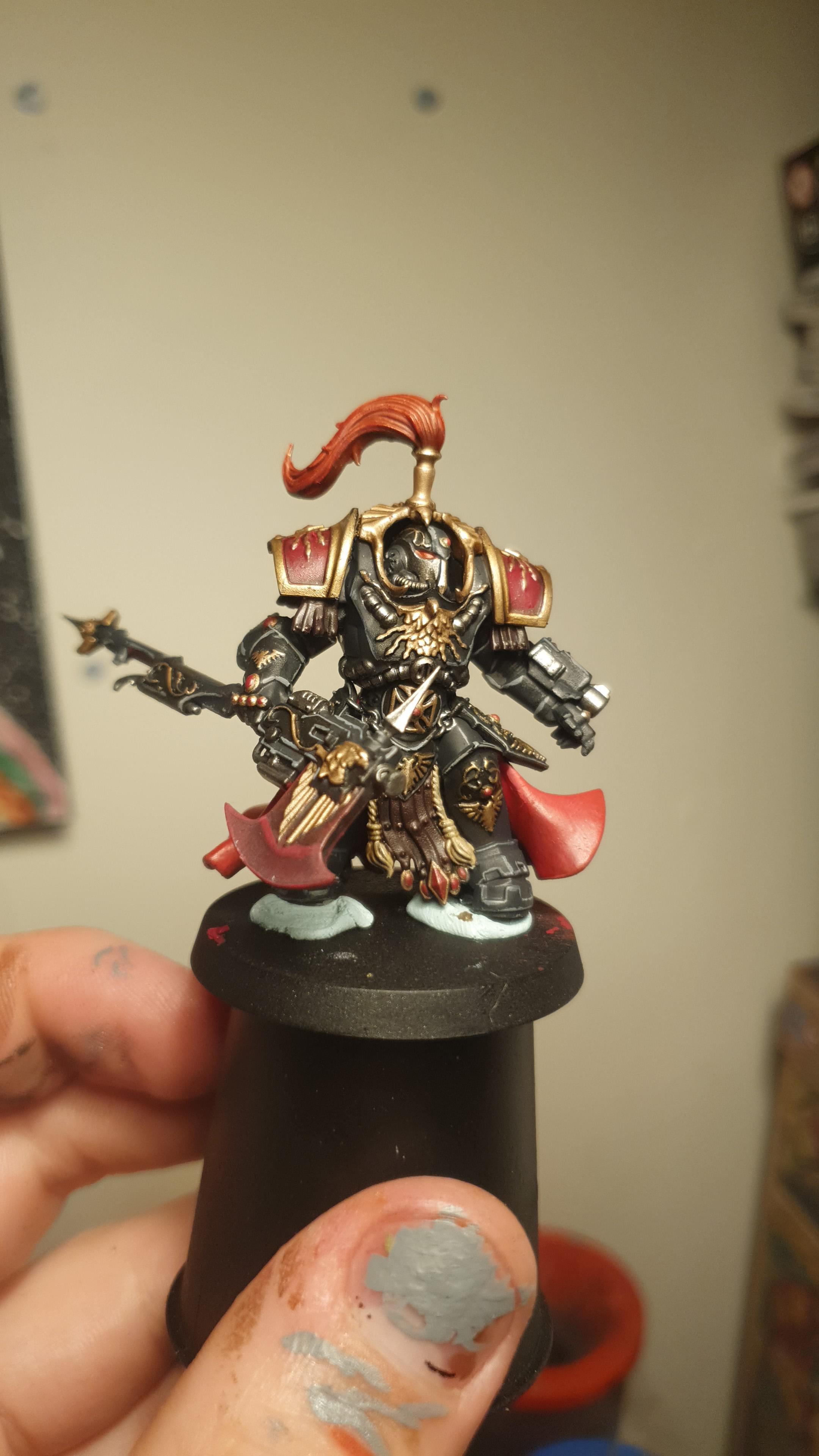 A hand holding a painted Warhammer miniature figure of a Shadowkeeper warrior with gold and red accents.