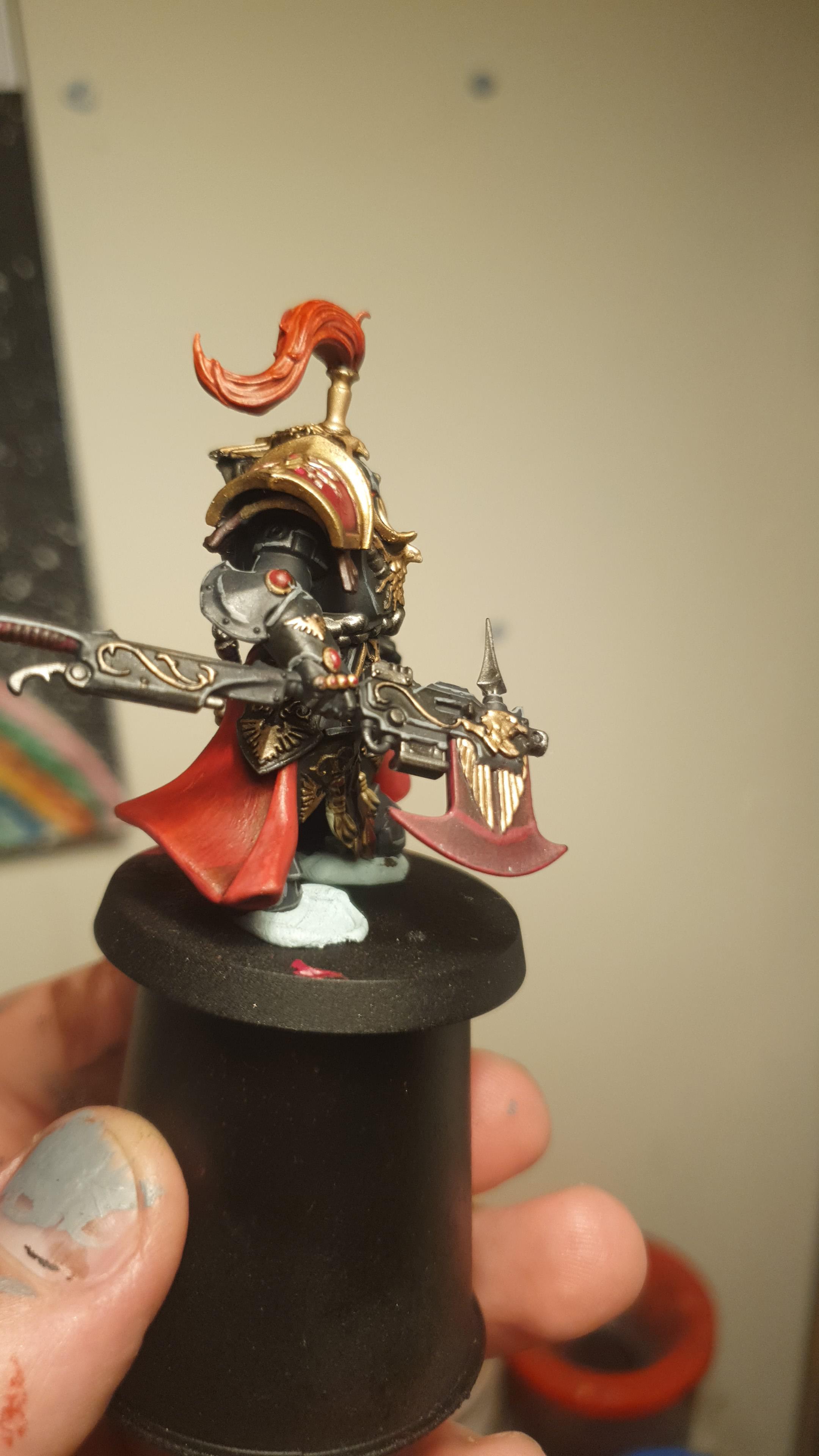 A hand holding a painted Warhammer miniature figure of a Shadowkeeper warrior with gold and red accents.