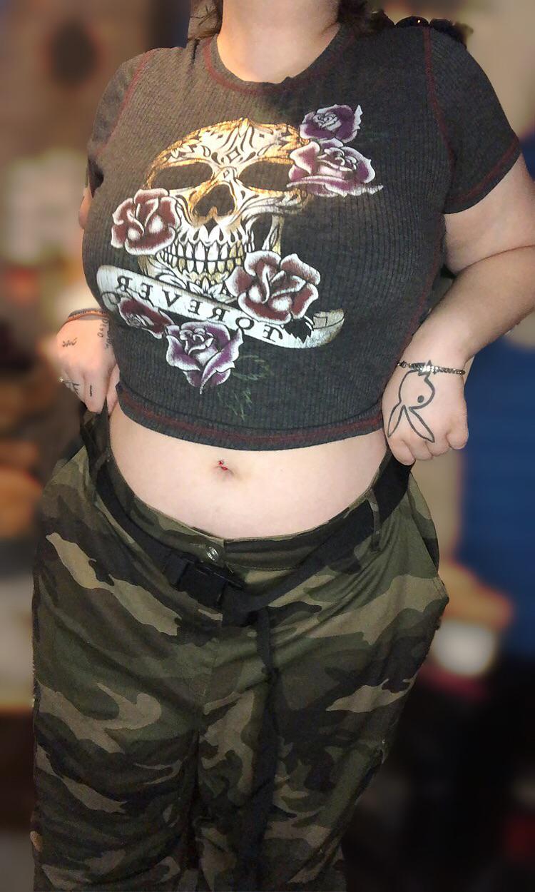 Curvy brunette woman in a cropped skull t-shirt and camouflage pants showing her midriff and belly piercing