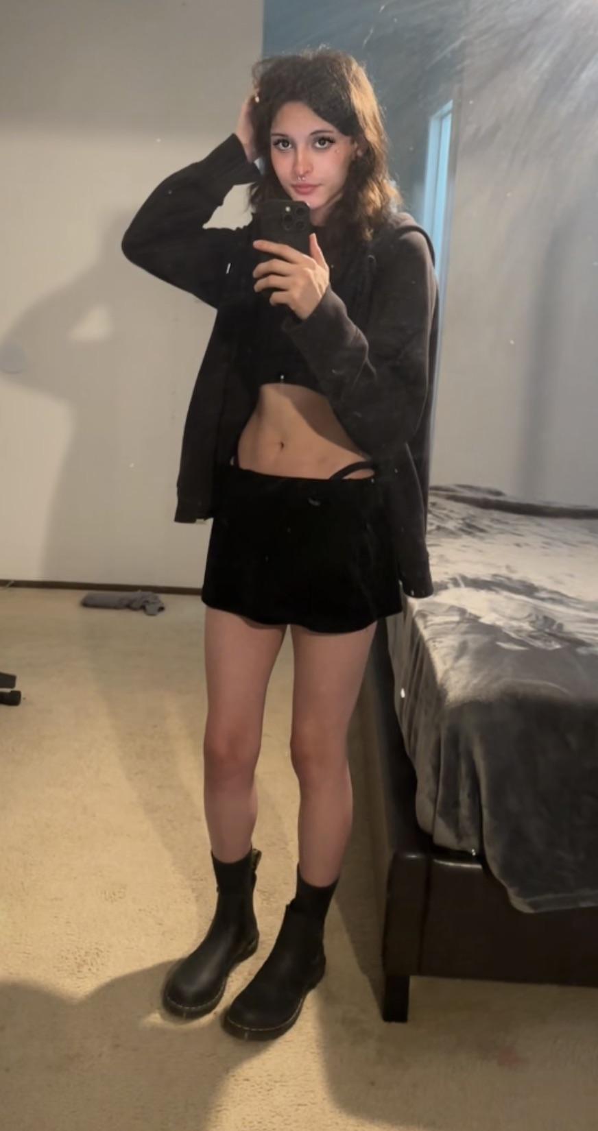 Androgynous hottie in a black cropped top, blazer, mini skirt, and ankle boots posing for mirror selfies.