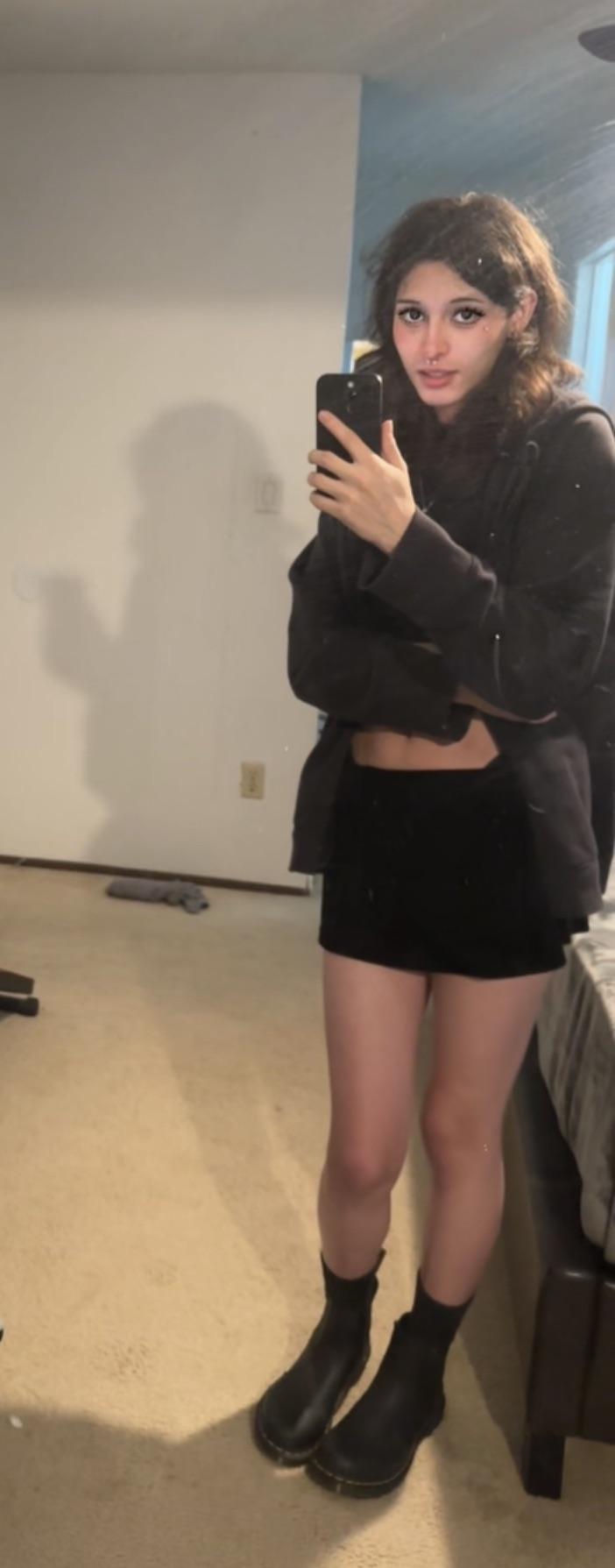 Androgynous hottie in a black cropped top, blazer, mini skirt, and ankle boots posing for mirror selfies.