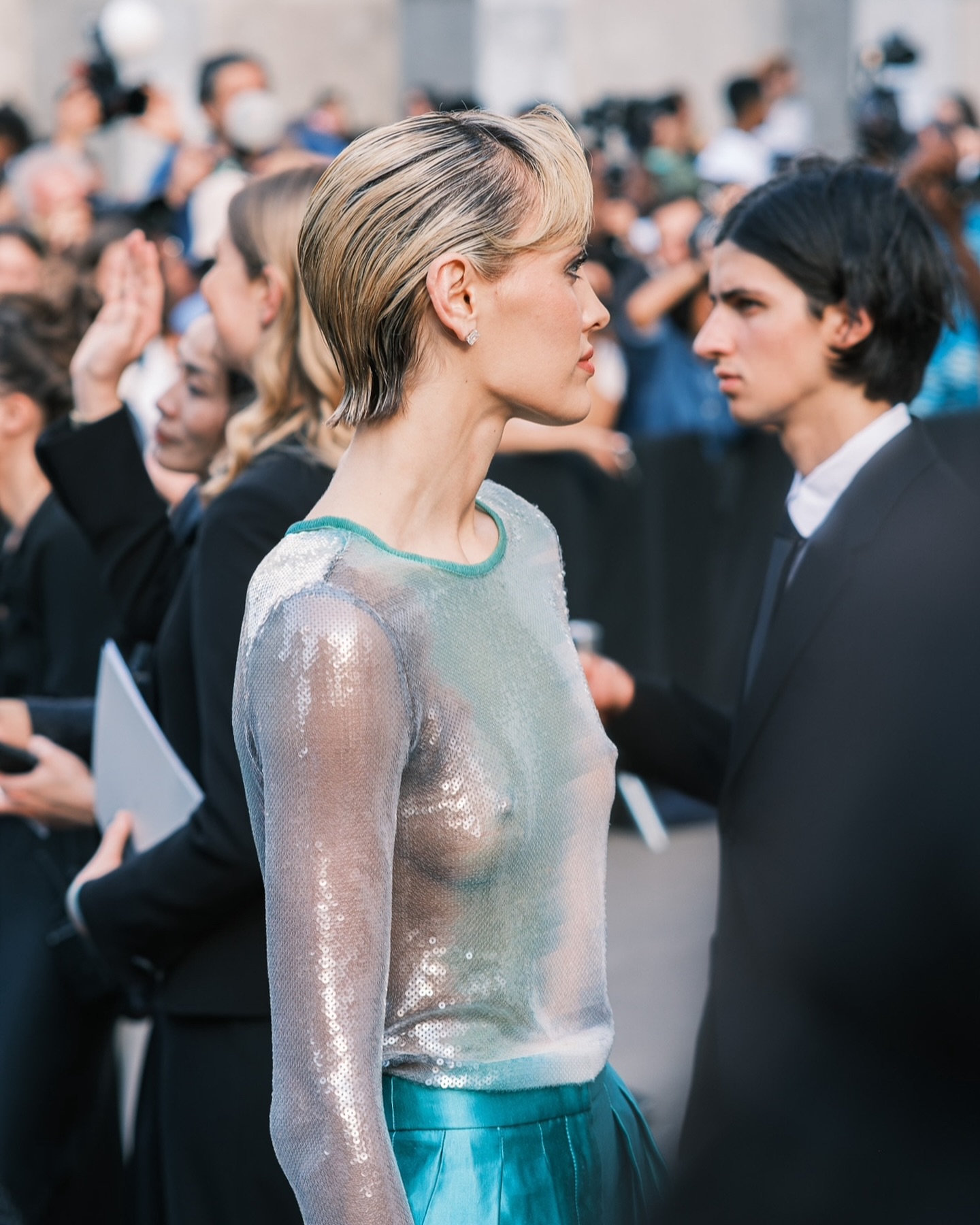 Wallis Day in a sheer silver sequined top and teal skirt standing with a man in a black suit