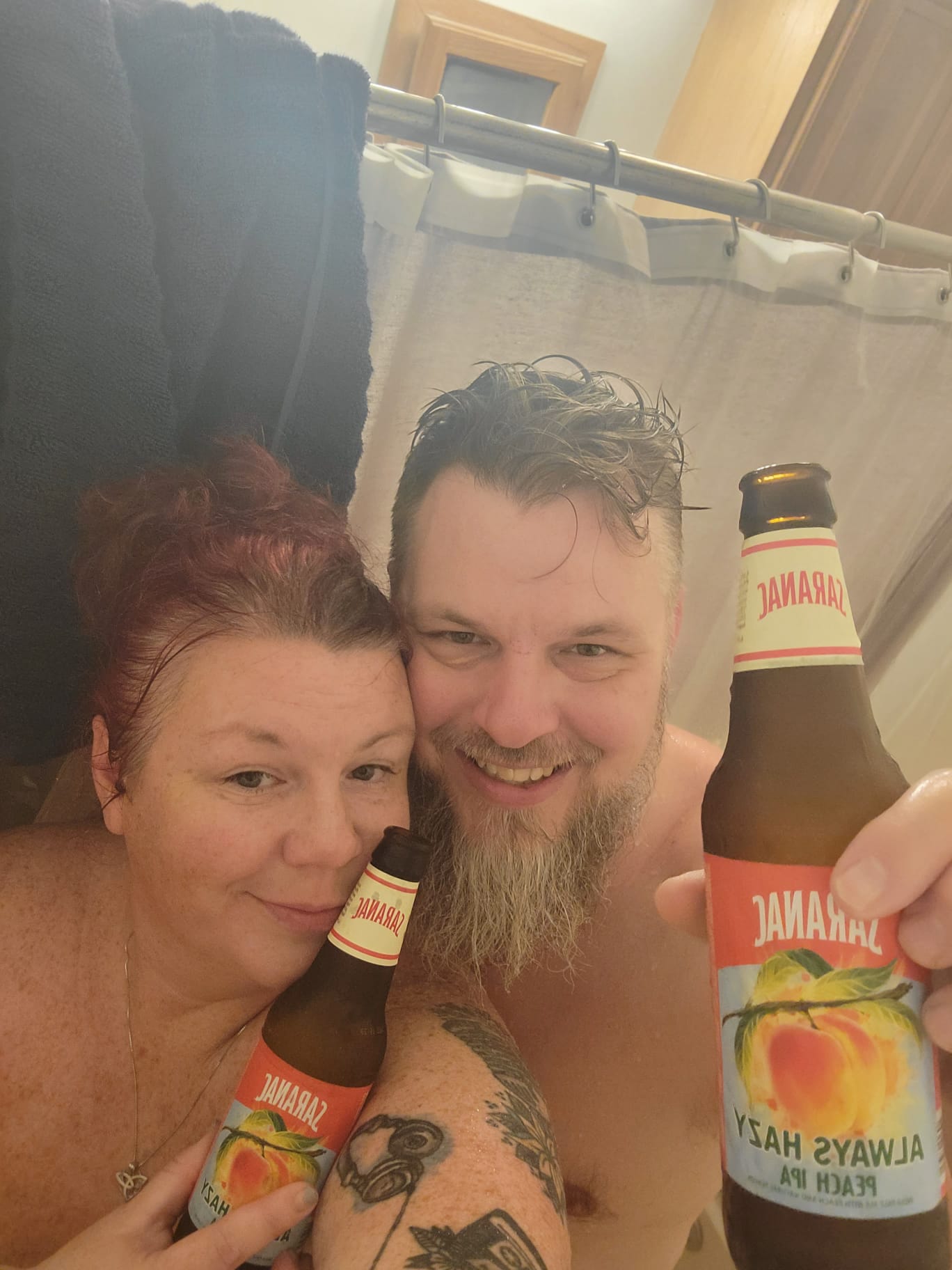 Naked redhead woman with tattoos and a man with a brown beard posing for a shower beer selfie in a bathtub
