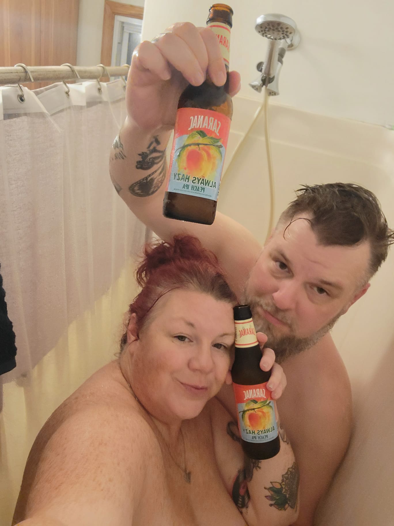 Naked redhead woman with tattoos and a man with a brown beard posing for a shower beer selfie in a bathtub