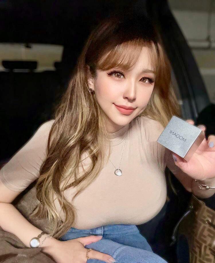 Busty Asian woman with long brown hair and bangs in a beige tight top and blue jeans posing with MADOM makeup in a car