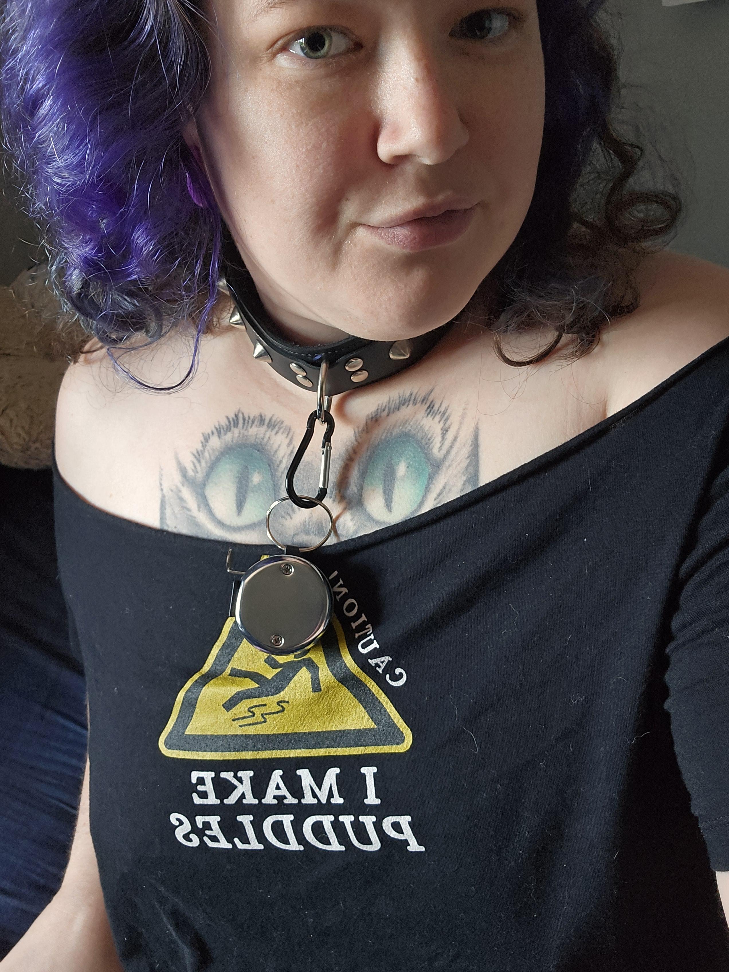 Busty white woman with purple curly hair wearing a spiked collar and nipple clamps, lifting her black shirt