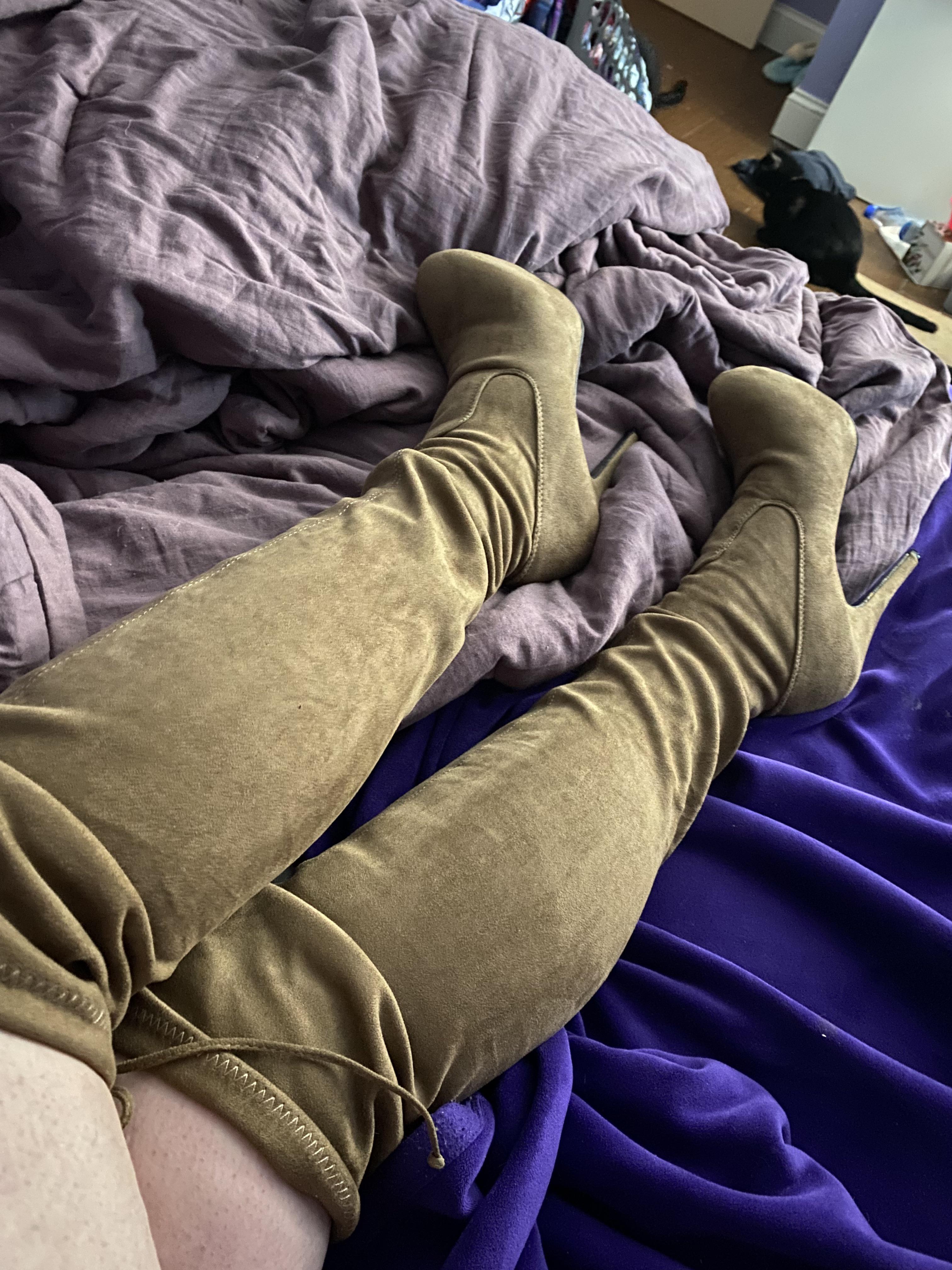 A person with fair skin wearing brown heeled thigh high boots lounging on a bed with a stuffed animal