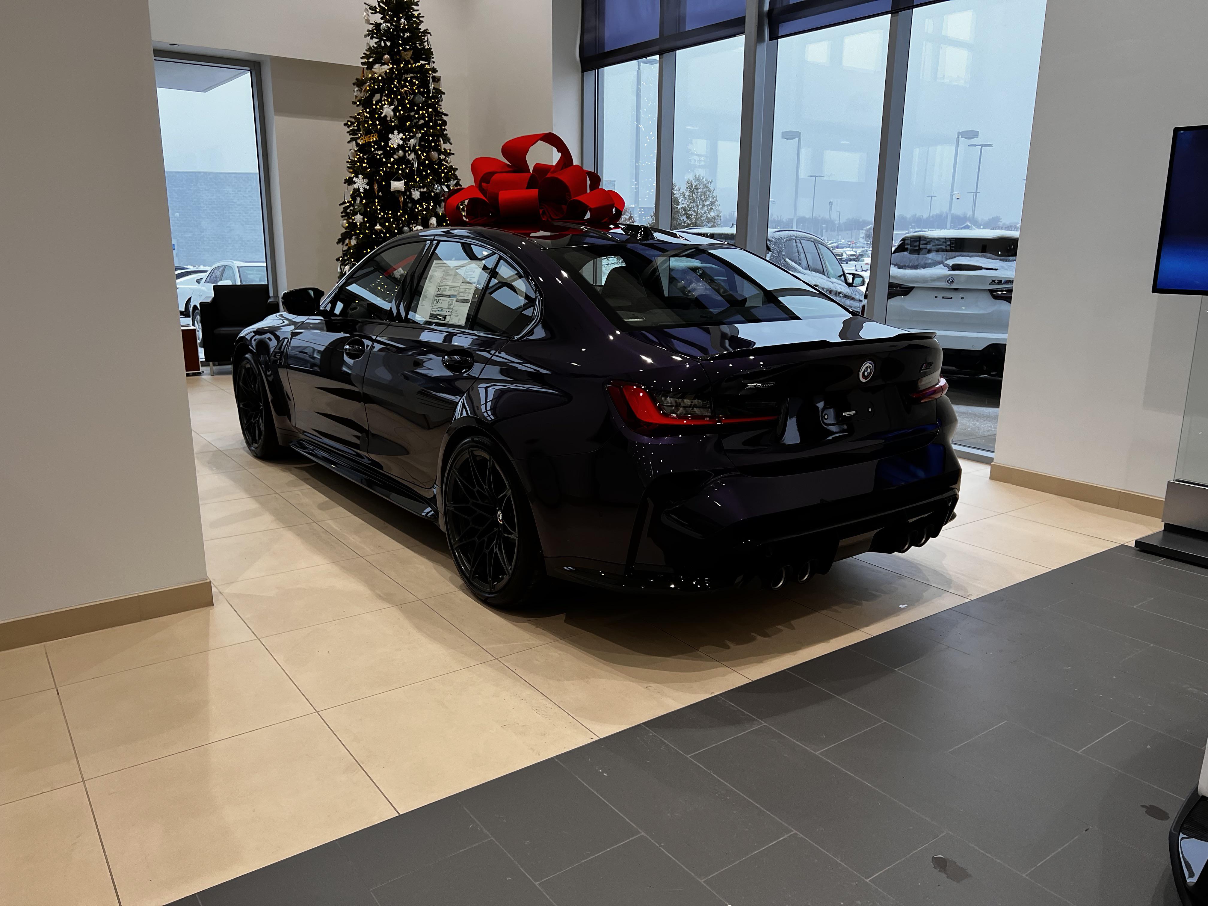 Dark purple BMW car with a large red bow parked next to a Christmas tree in a dealership