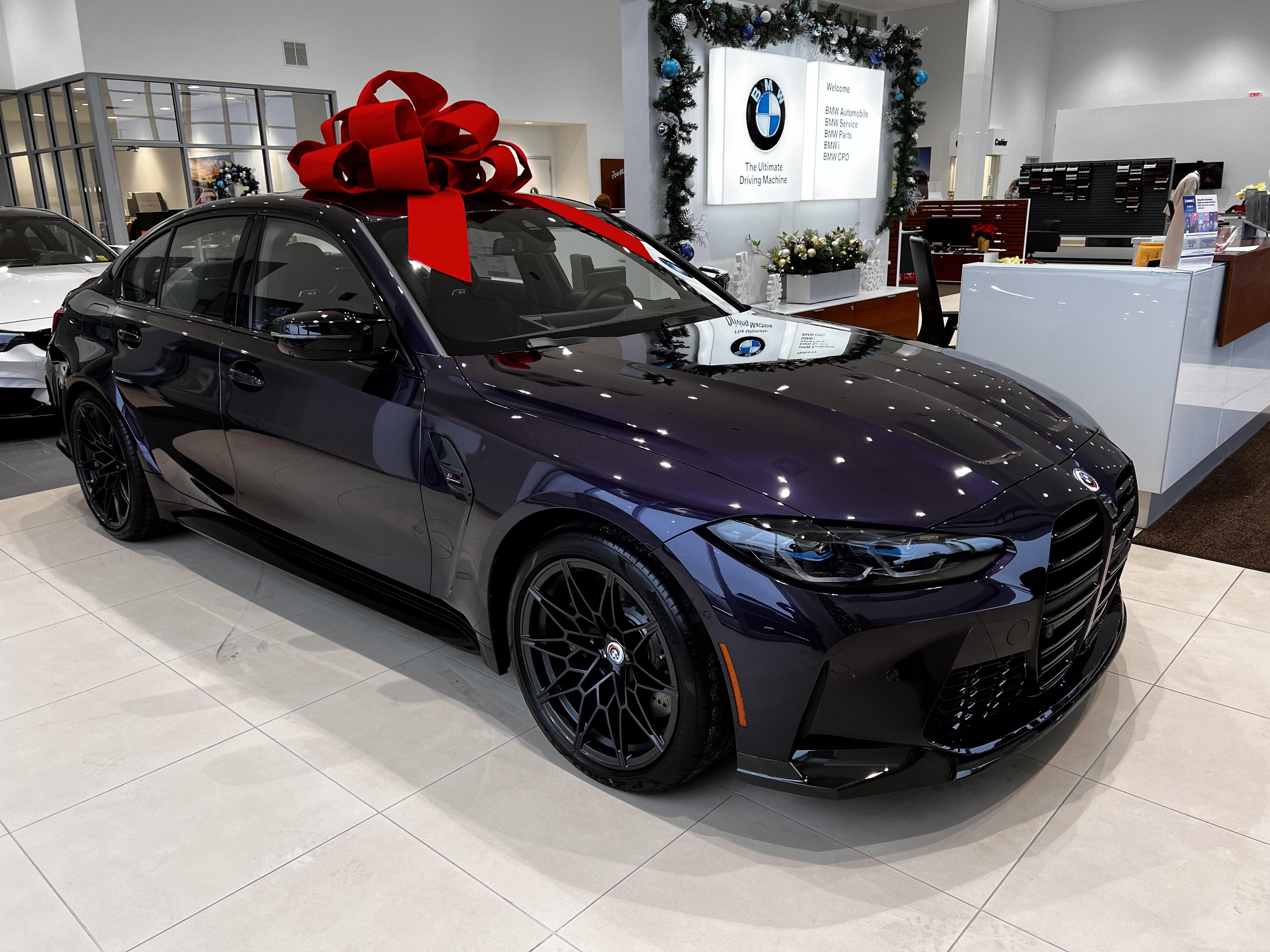 Dark purple BMW car with a large red bow parked next to a Christmas tree in a dealership