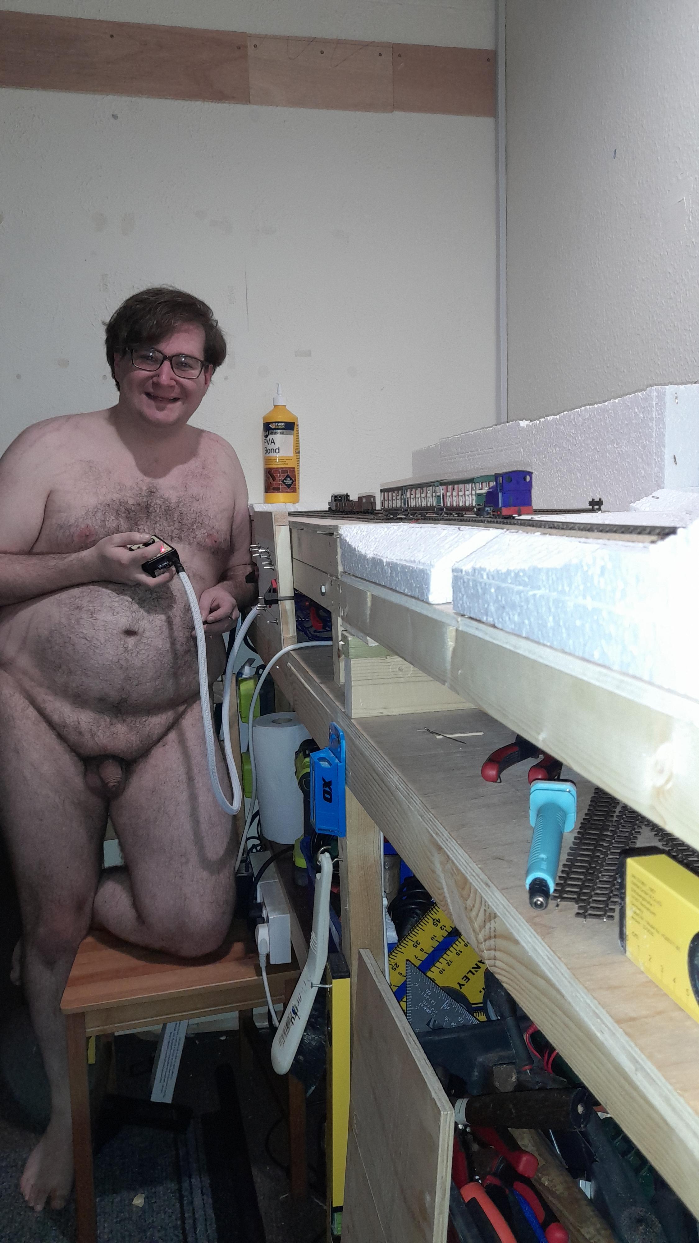 Hairy white bbw man posing nude in a workshop with a model train layout