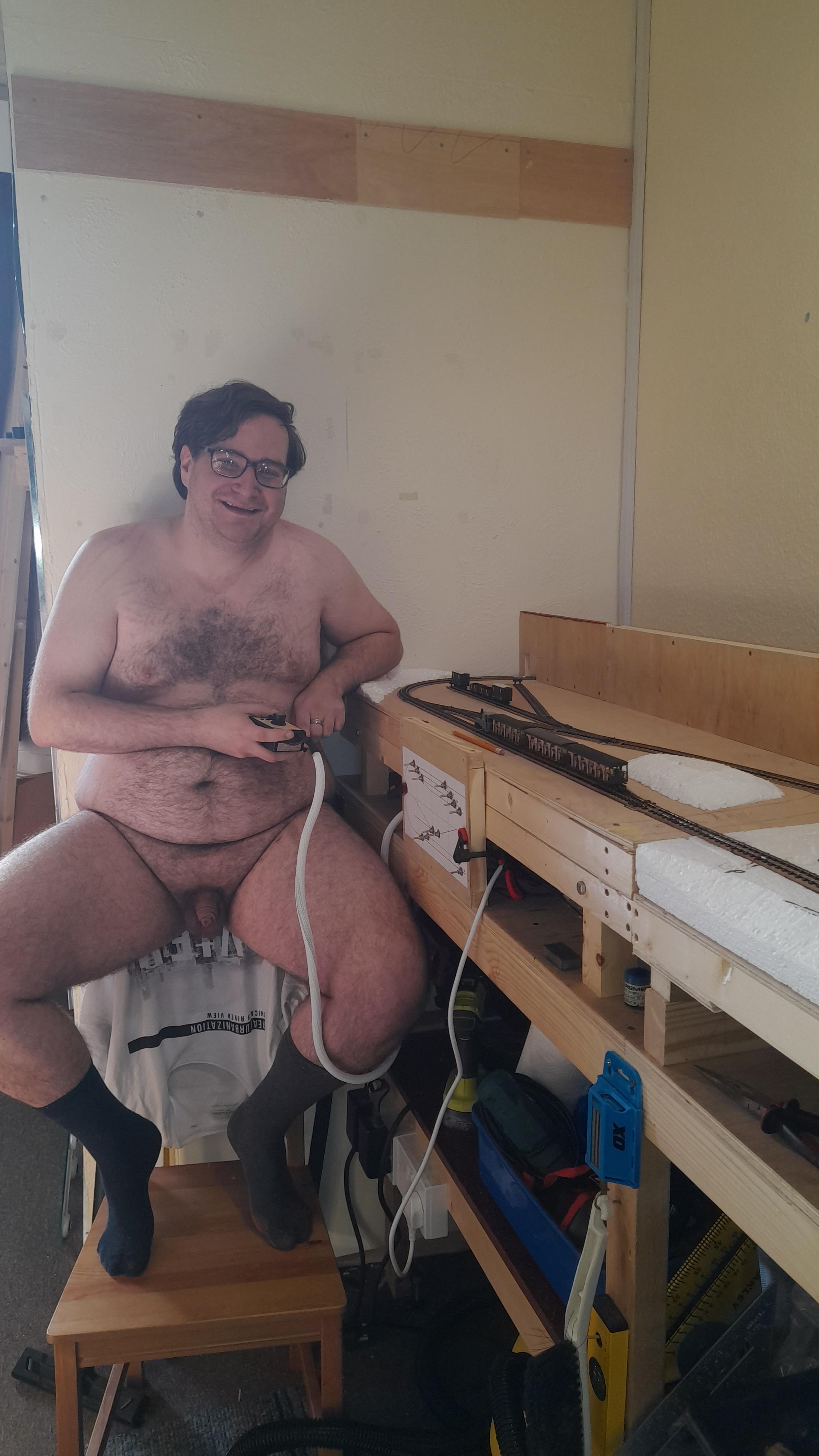 Hairy white bbw man posing nude in a workshop with a model train layout