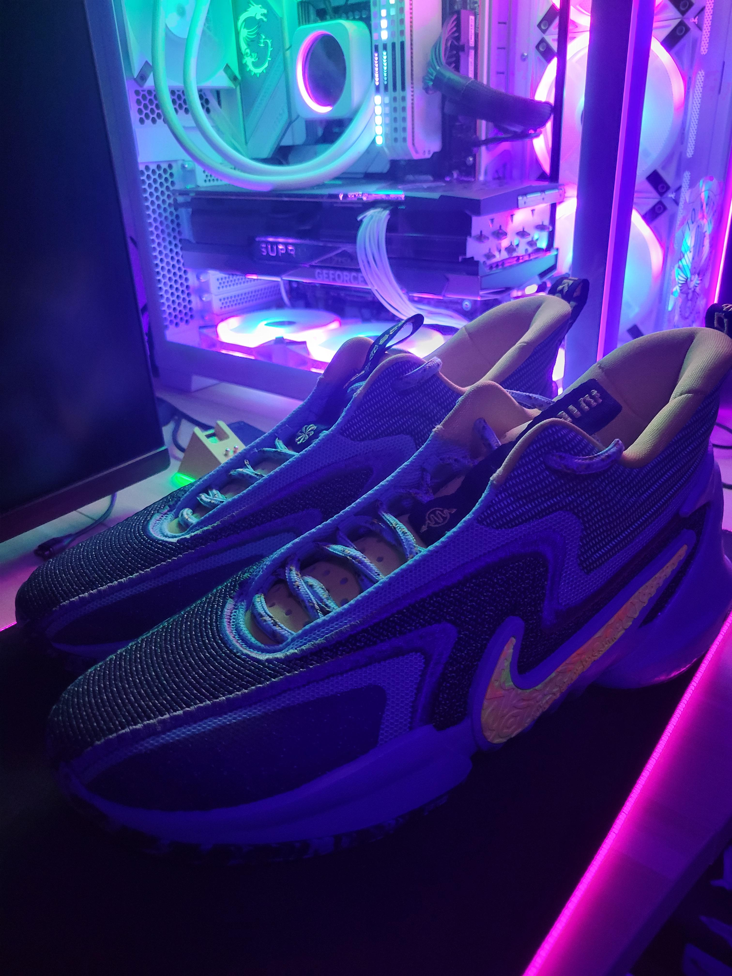 Blue and yellow Nike sneakers in a close-up shot in front of a glowing gaming PC desk setup.