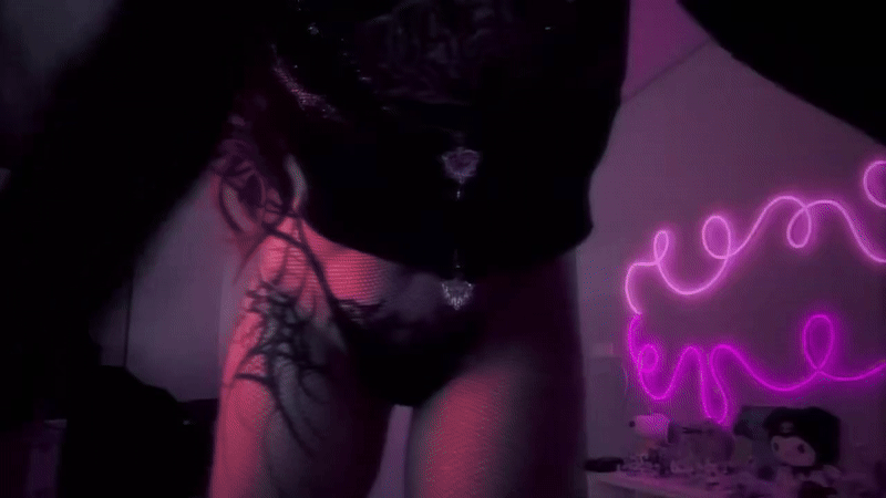 Goth girl with tattoos in a black crop top and thong posing under pink neon lights
