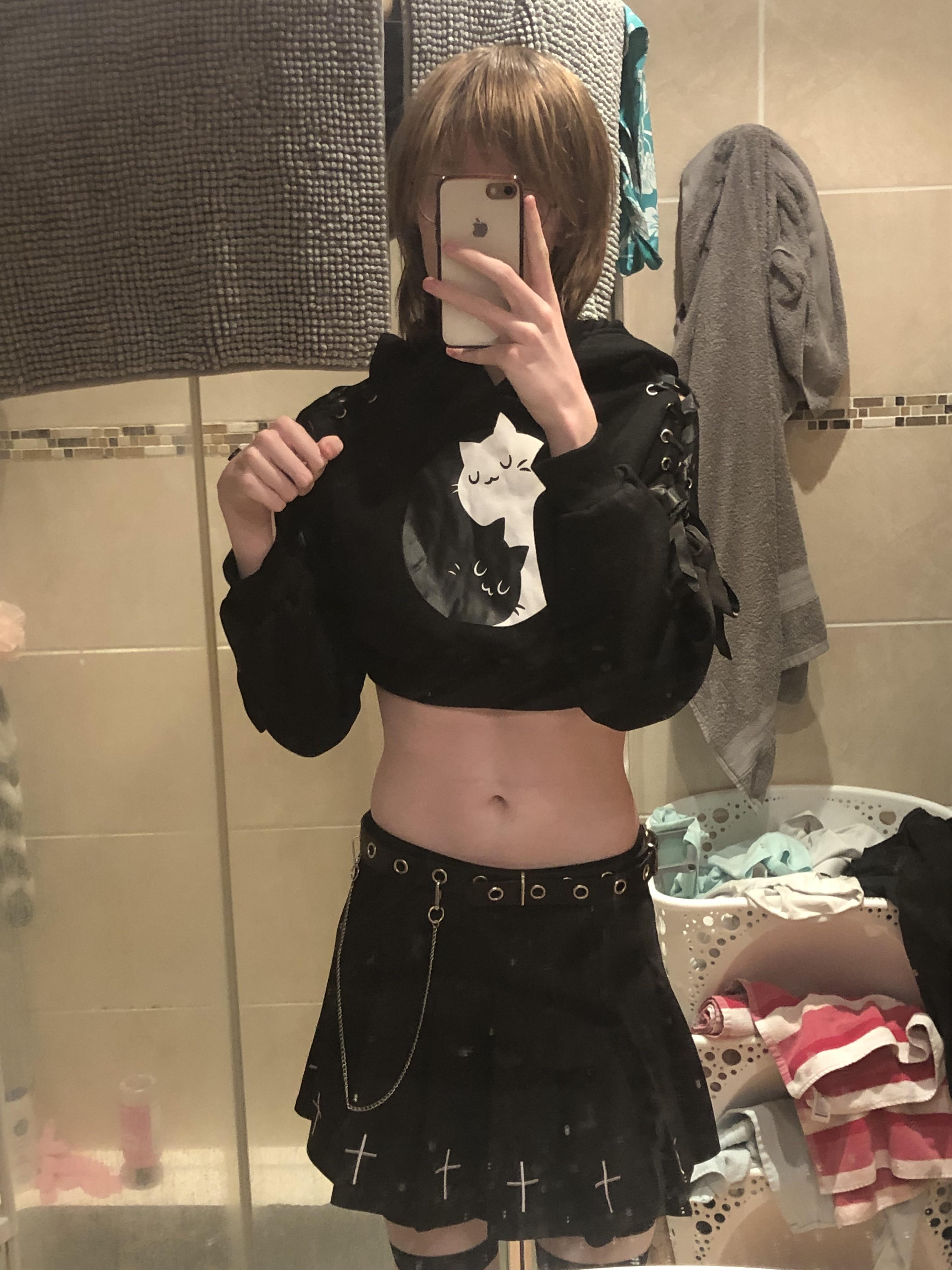 A slender femboy with short blonde hair taking a mirror selfie in a black cropped top and pleated mini skirt.