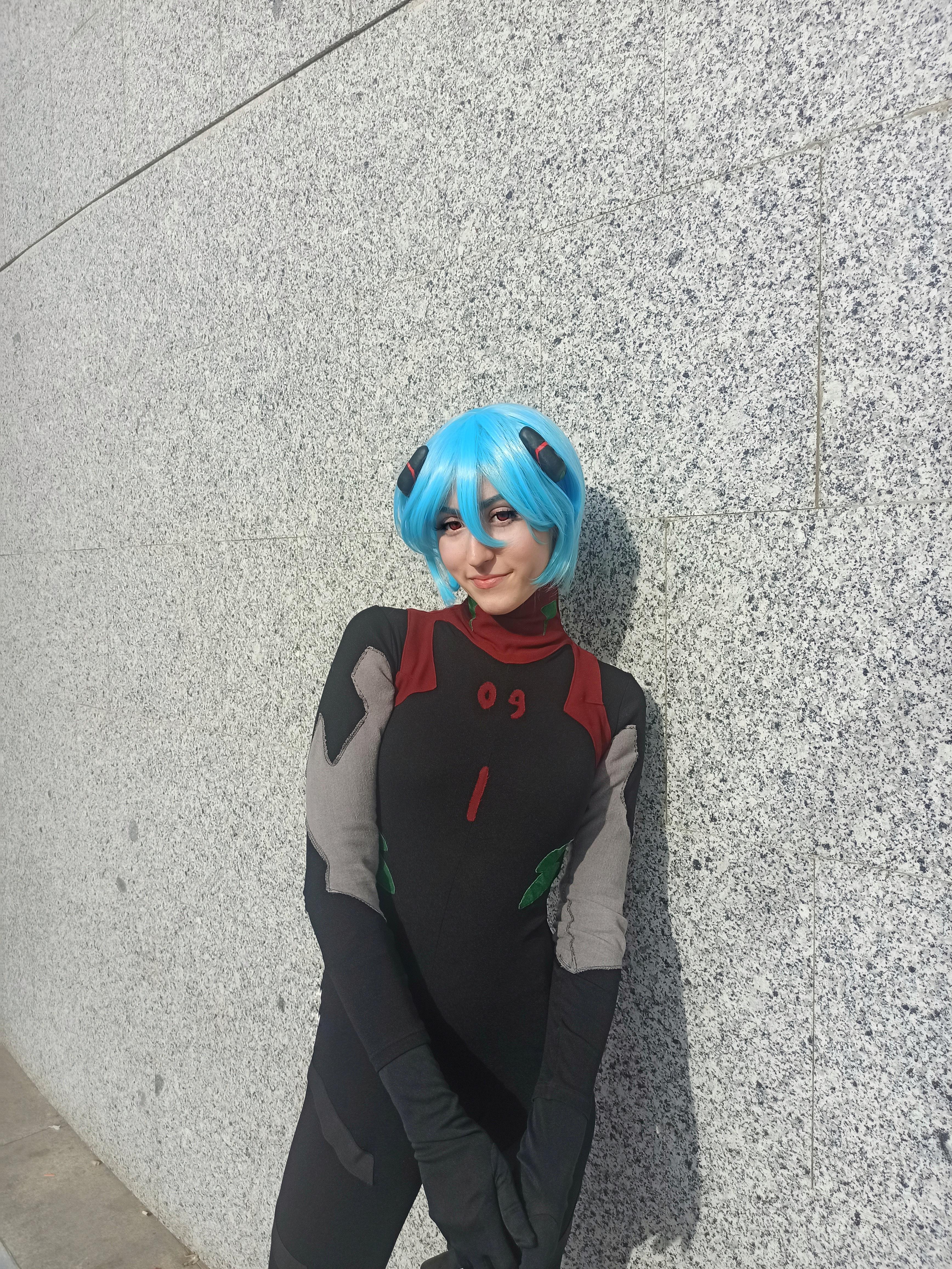Rei Ayanami cosplay in a black plugsuit with a short blue wig posing against a grey stone wall