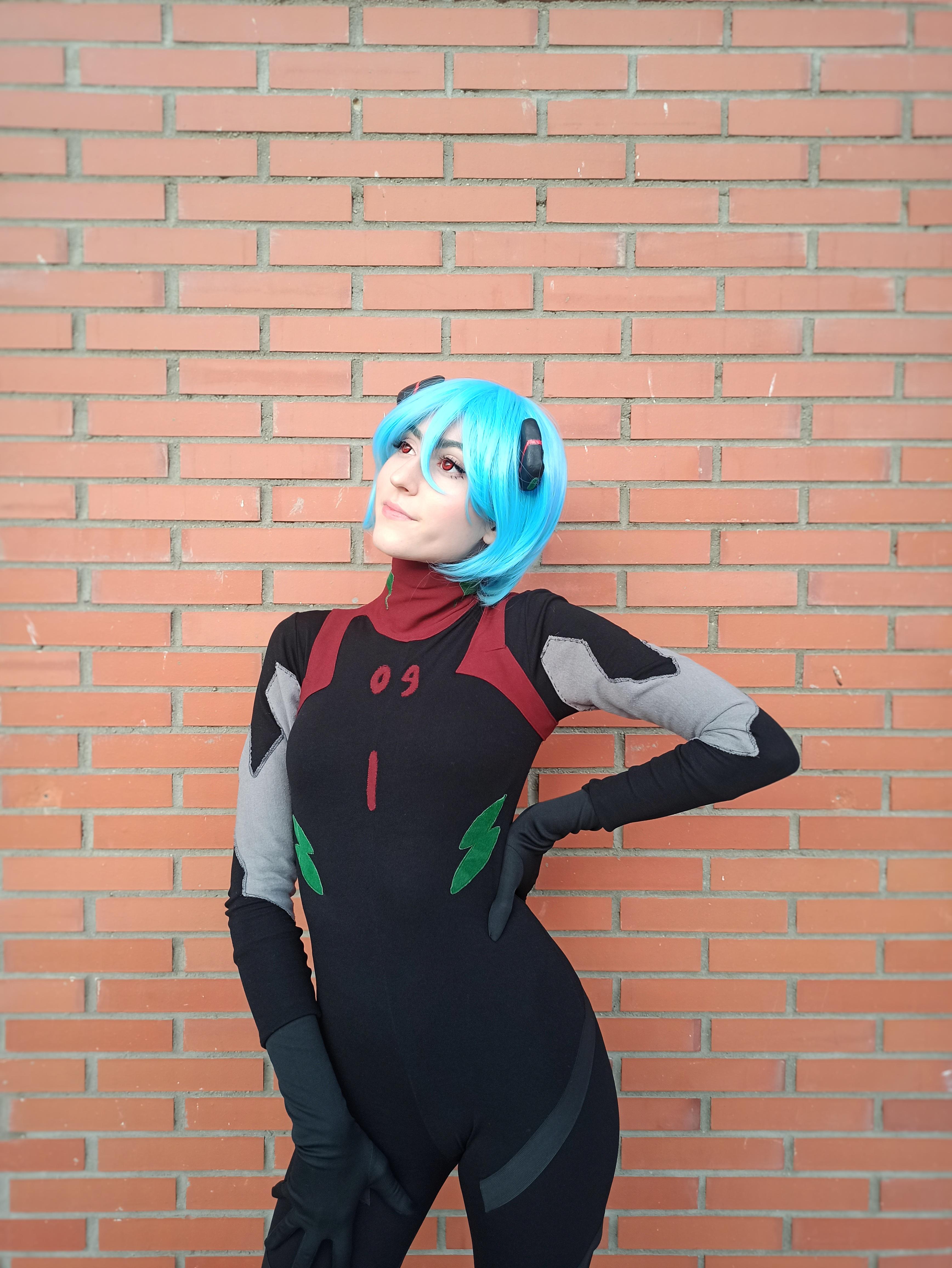 Rei Ayanami cosplay in a black plugsuit with a short blue wig posing against a grey stone wall