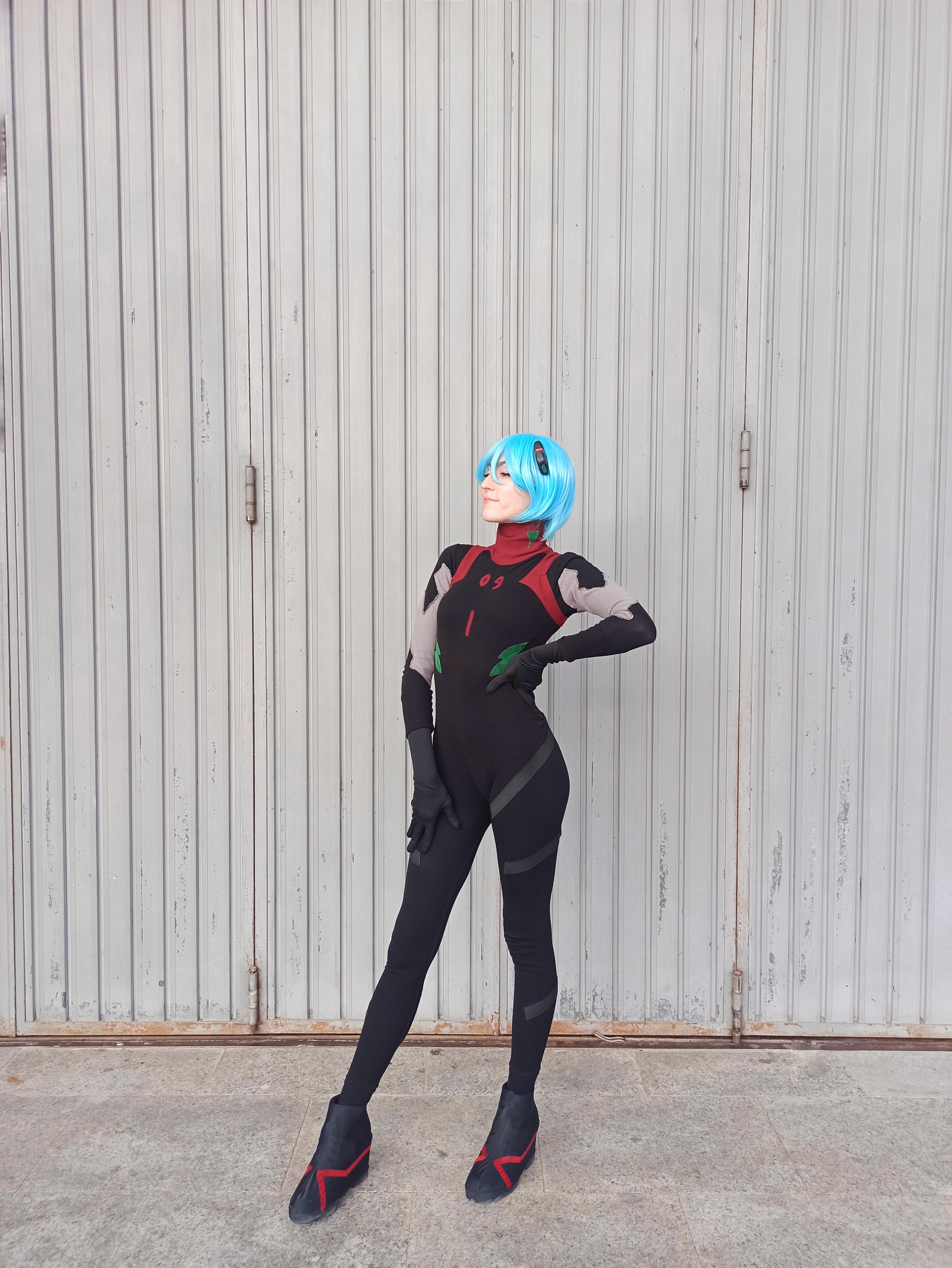 Rei Ayanami cosplay in a black plugsuit with a short blue wig posing against a grey stone wall