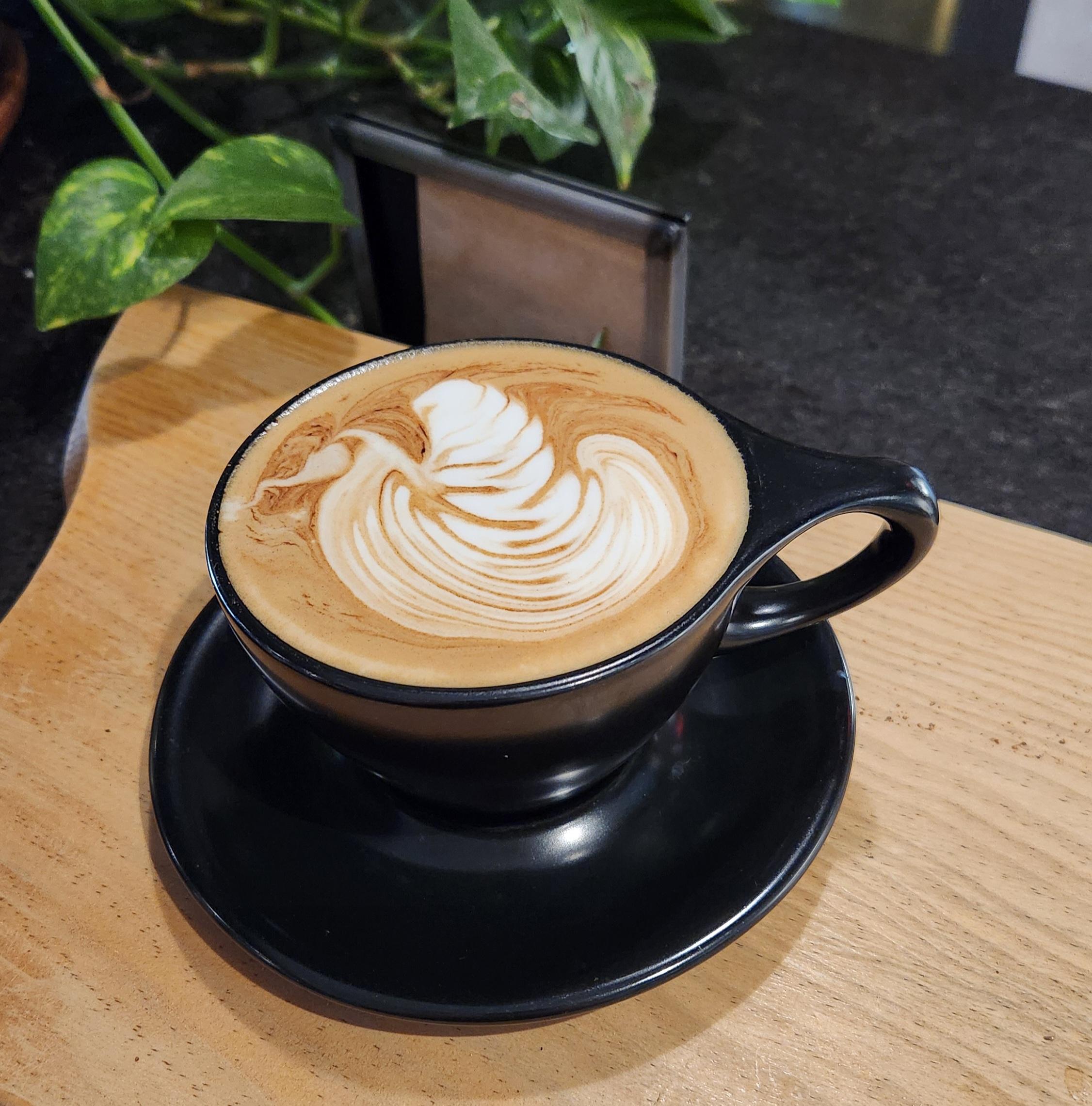 Latte art with a leaf-shaped foam design in a black cup on a wooden surface with ivy houseplants