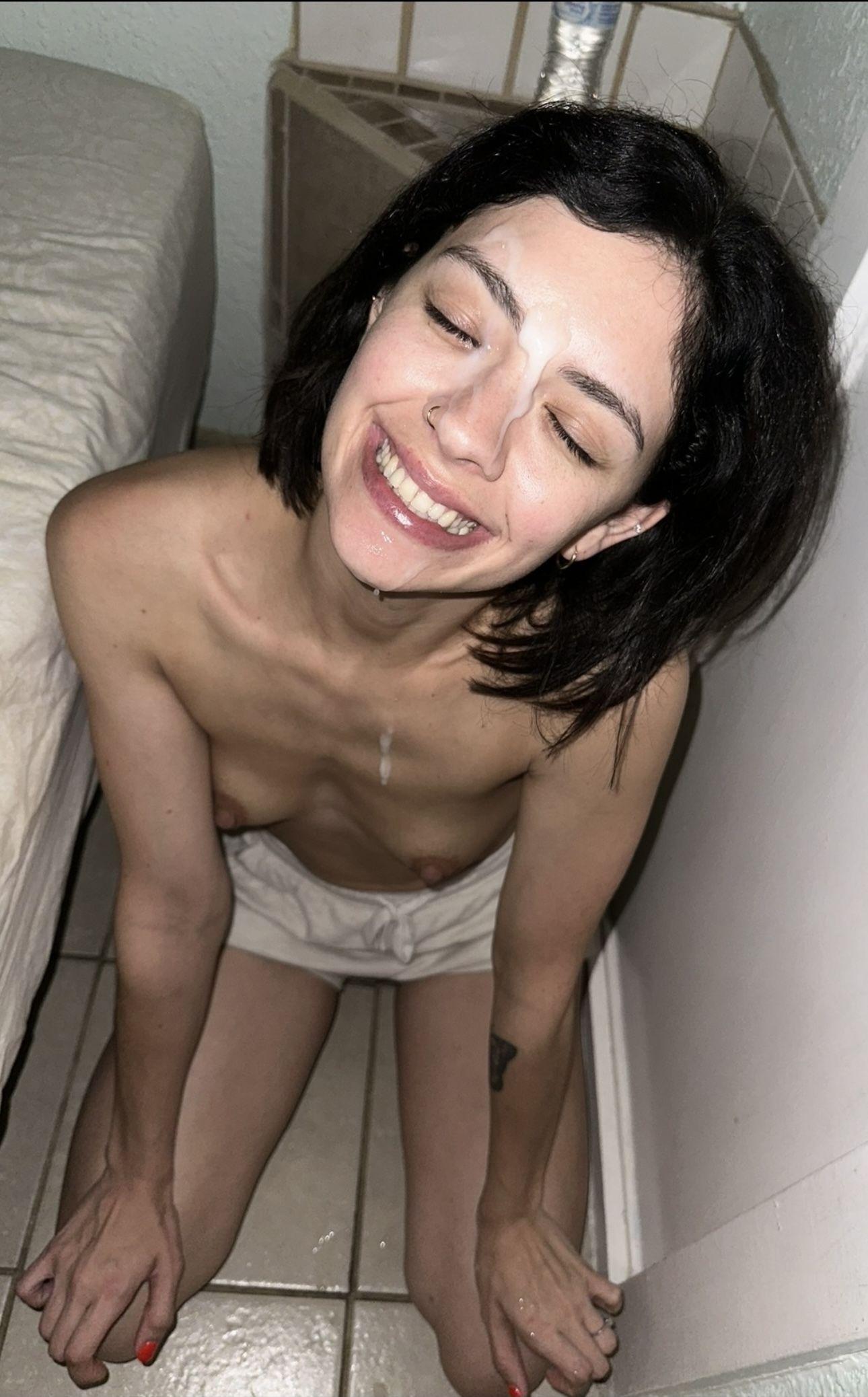 Smiling white woman with short dark hair, nose ring, and tattoo, topless in white shorts with a facial cumshot