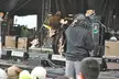 Kanye West in a baseball cap and ripped jeans performing on an outdoor concert stage with crew and a crowd.