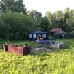 Campsite in a grassy field with sofas, a fire pit, a blue gazebo, and tents surrounded by trees.