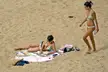 Two women in patterned bikinis on a sandy beach, one lying on a towel and the other standing with a bottle.