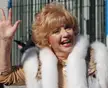Ruta Lee smiling and waving, wearing a white fur vest and red lipstick.