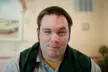 A man with a skeptical, weird face wearing a grey shirt and dark green jacket, a funny profile picture meme.