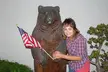 Dom, a country girl with brown hair and a plaid shirt, smiling next to a bear statue with an American flag